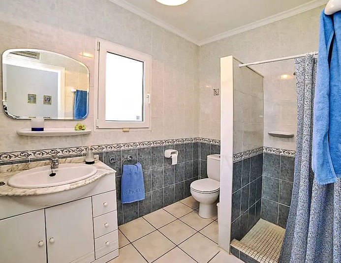 Baño funcional con azulejos en dos tonos, encimera de mármol y ducha de obra. Dispone de ventana de PVC blanca para ventilación natural y un mueble de lavabo con almacenaje.