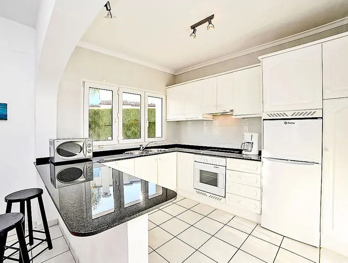 Cocina equipada con armarios blancos, encimera de granito negro y barra americana. Dispone de electrodomésticos integrados, suelos de baldosa y luz natural desde una ventana frontal.