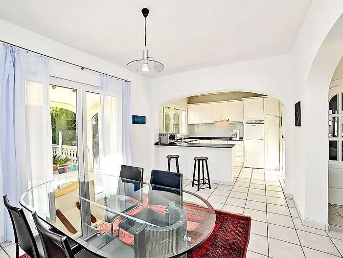 Comedor luminoso con mesa de cristal y alfombra roja, conectado a una cocina blanca con barra americana a través de un arco arquitectónico.