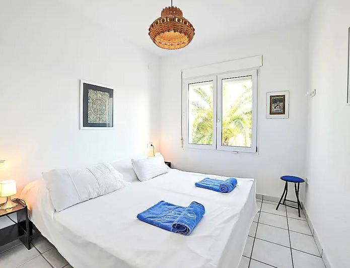 Dormitorio luminoso con paredes blancas, suelos de baldosa y una ventana con vistas a las palmeras. Incluye una lámpara de mimbre y mobiliario funcional.