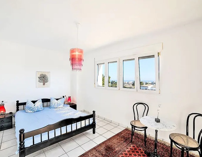 Dormitorio luminoso con suelos de baldosa blanca, cama de madera oscura y zona de estar con mesa de mármol. Las ventanas ofrecen vistas despejadas hacia la costa de Dénia.