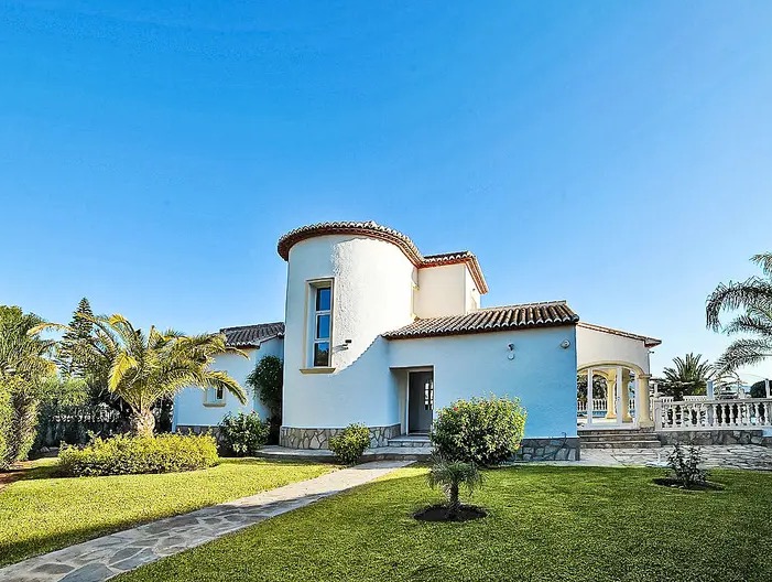 Fachada exterior de la villa con torre circular, paredes blancas y tejado de terracota. Incluye un jardín cuidado con césped y palmeras bajo un cielo despejado.