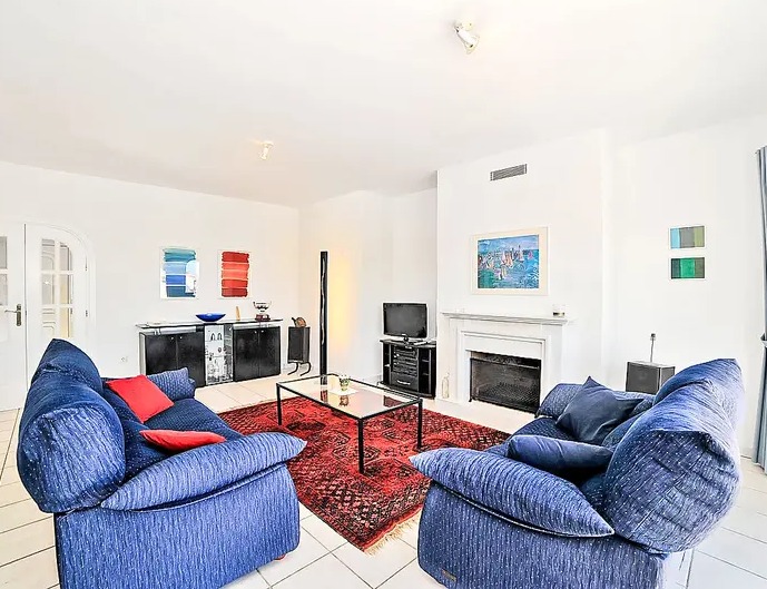 Salón luminoso con chimenea blanca, sofás azules, alfombra roja de estilo oriental y suelos de baldosa clara que aportan amplitud.