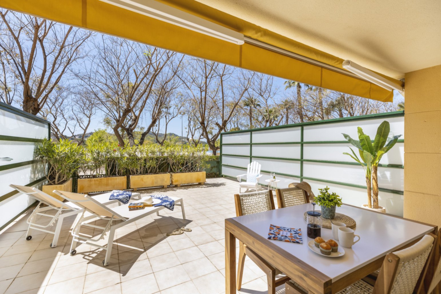 Amplia terraza de baldosas con toldo amarillo, zona de comedor exterior y tumbonas blancas, protegida por paneles de privacidad y rodeada de árboles.