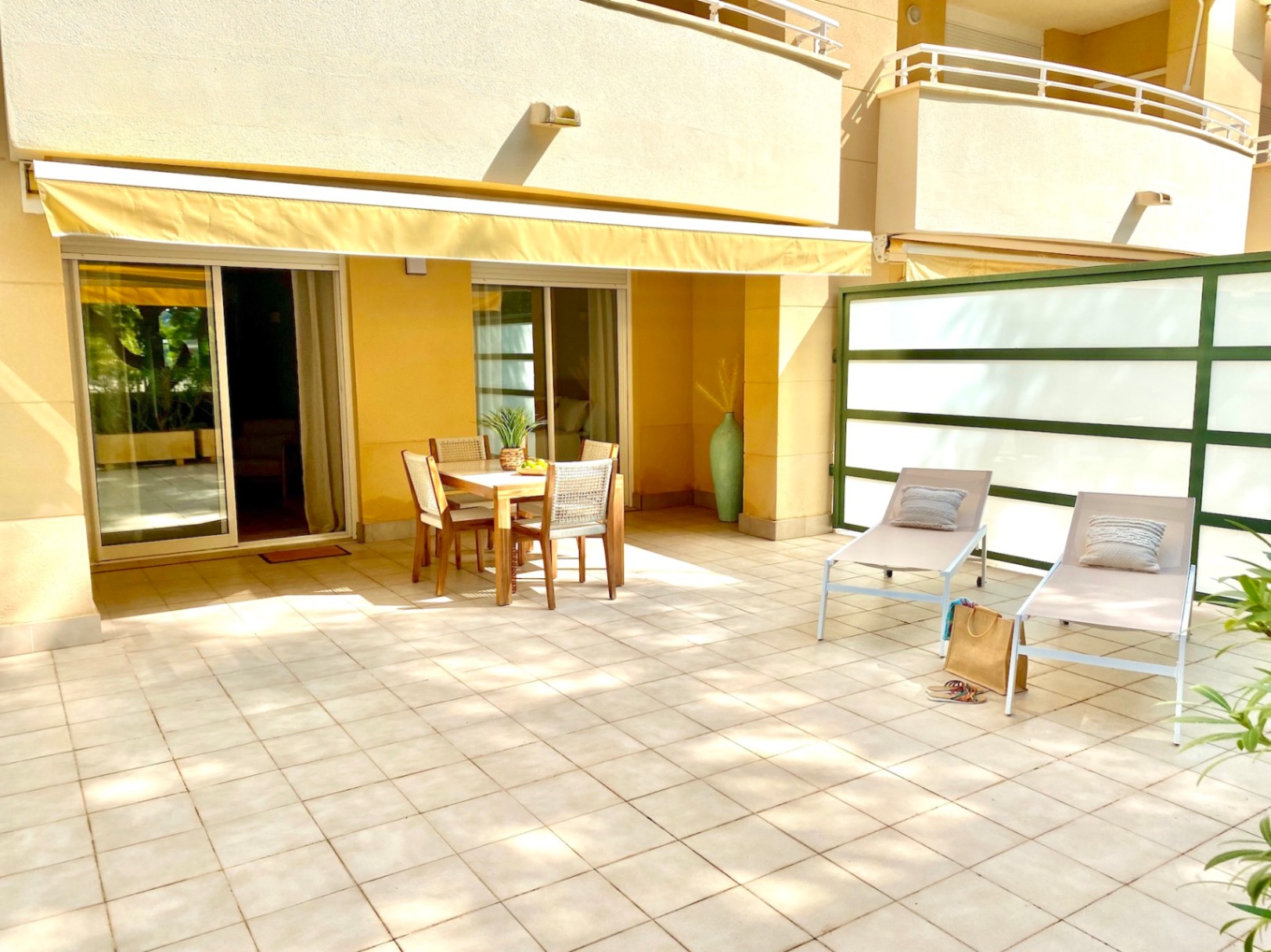 Amplia terraza pavimentada con comedor de madera bajo un toldo amarillo extensible y dos tumbonas junto a un panel separador translúcido.