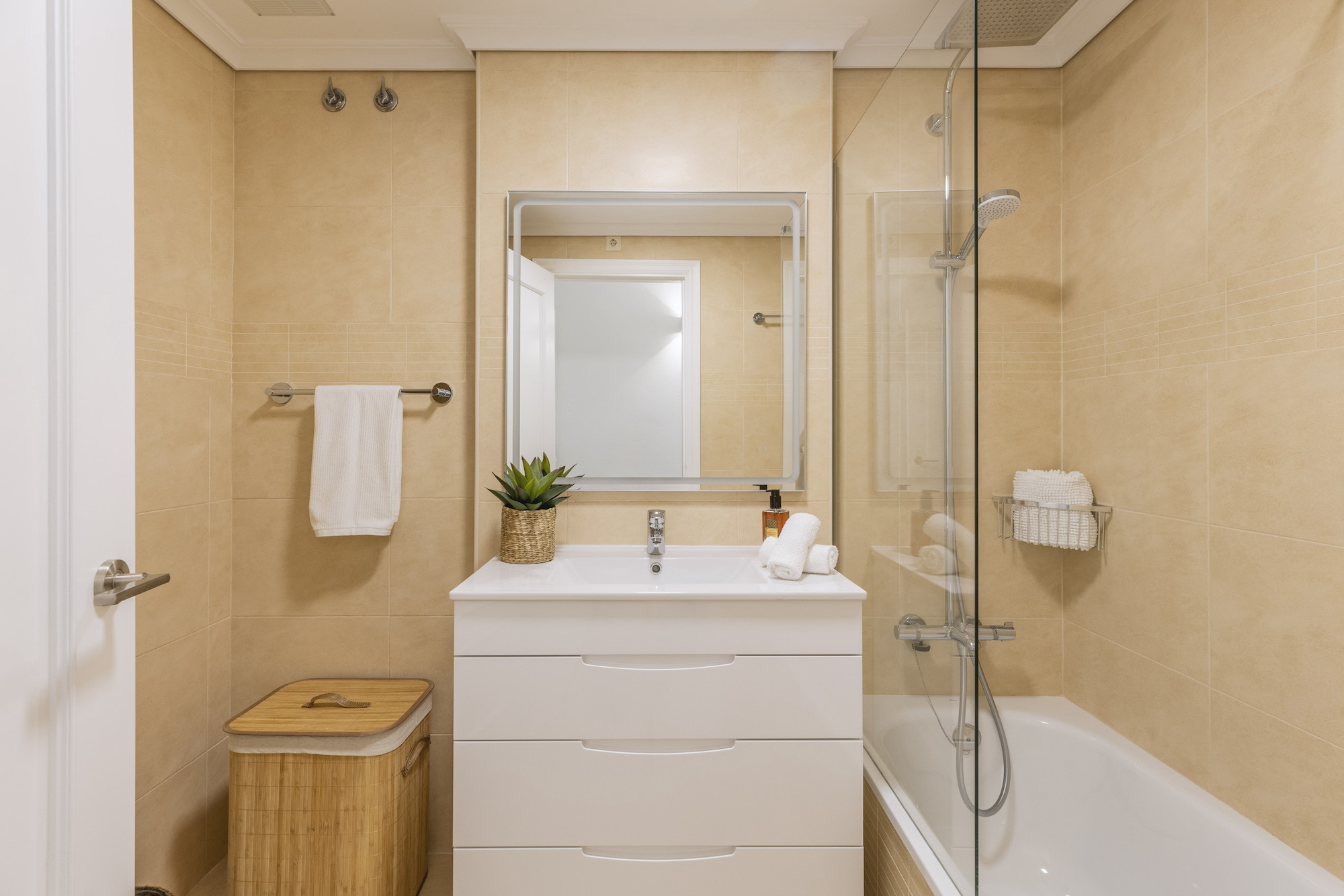 Baño moderno con azulejos en tonos beige, mueble de lavabo blanco con tres cajones, espejo retroiluminado y bañera con mampara de cristal.