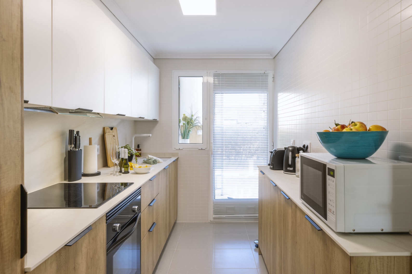 Cocina lineal moderna con armarios inferiores de efecto madera, muebles superiores blancos, electrodomésticos integrados y ventana con persianas.