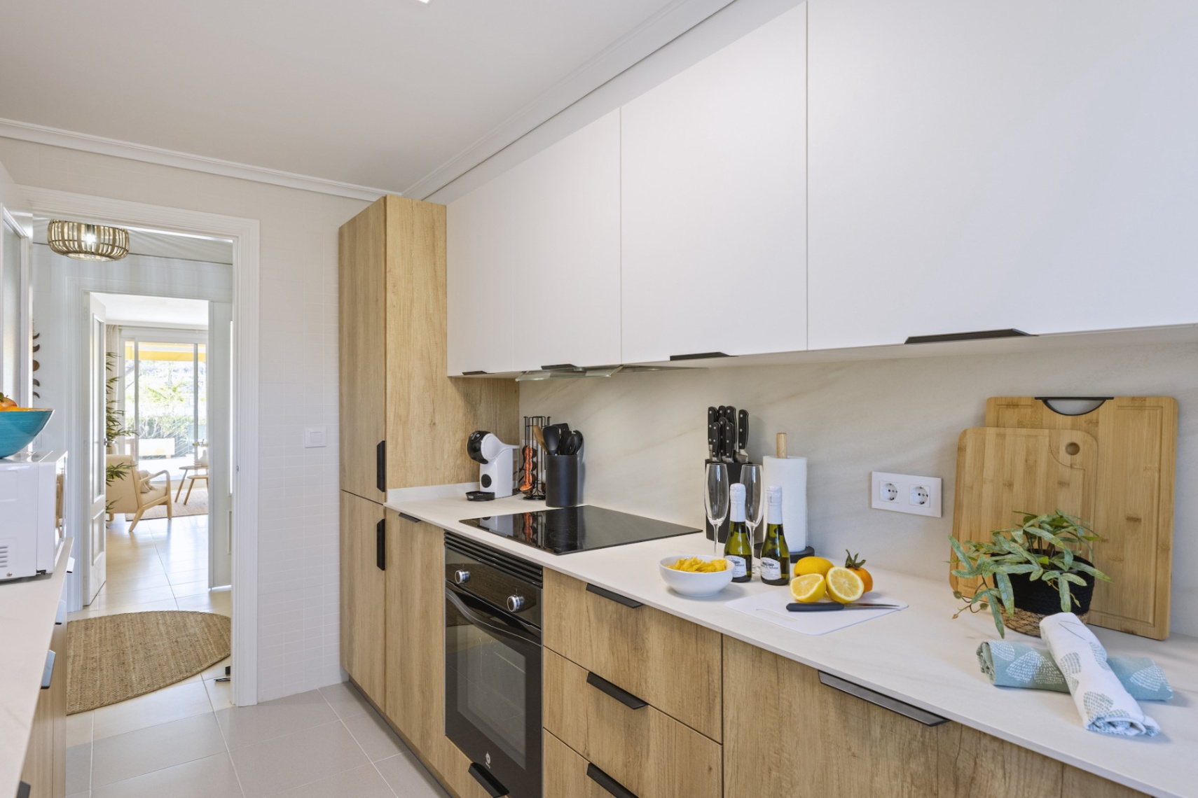 Cocina moderna con armarios bicolores en blanco y madera natural. Incluye placa de inducción, horno integrado y encimeras de piedra blanca con vista al salón.
