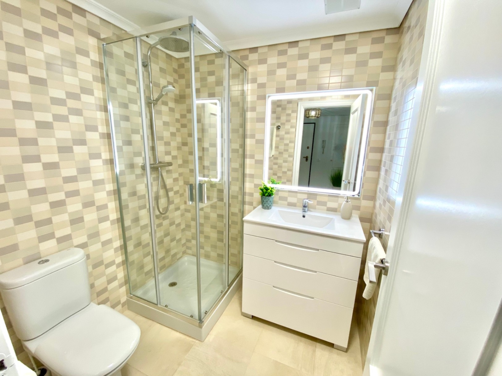 Cuarto de baño con ducha de esquina acristalada, lavabo sobre mueble blanco de tres cajones y espejo retroiluminado sobre paredes de mosaico beige.