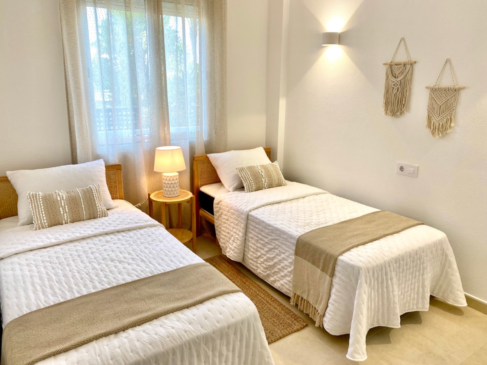 Dormitorio luminoso con dos camas individuales, cabeceros de mimbre, textiles en tonos neutros, decoración de macramé y una cálida iluminación natural y artificial.