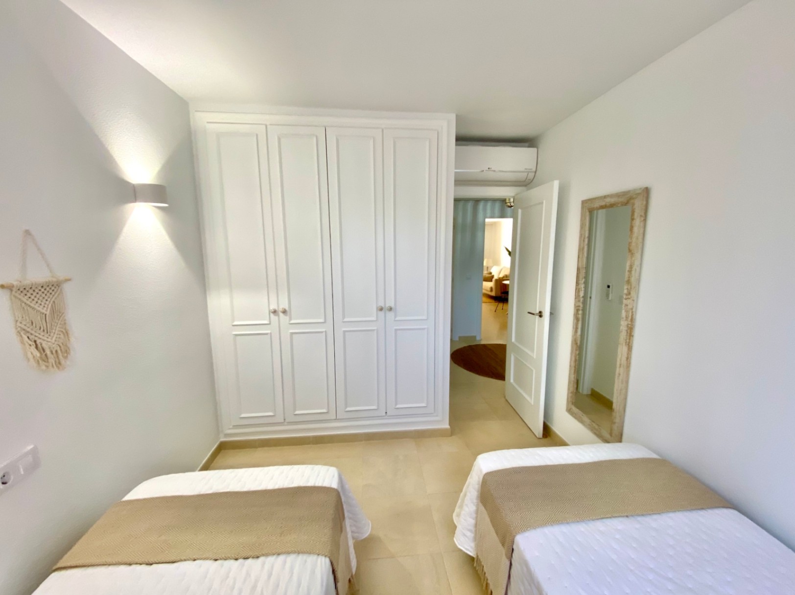 Dormitorio luminoso con dos camas individuales, un amplio armario empotrado blanco y un espejo con marco de madera rústica sobre suelos cerámicos.