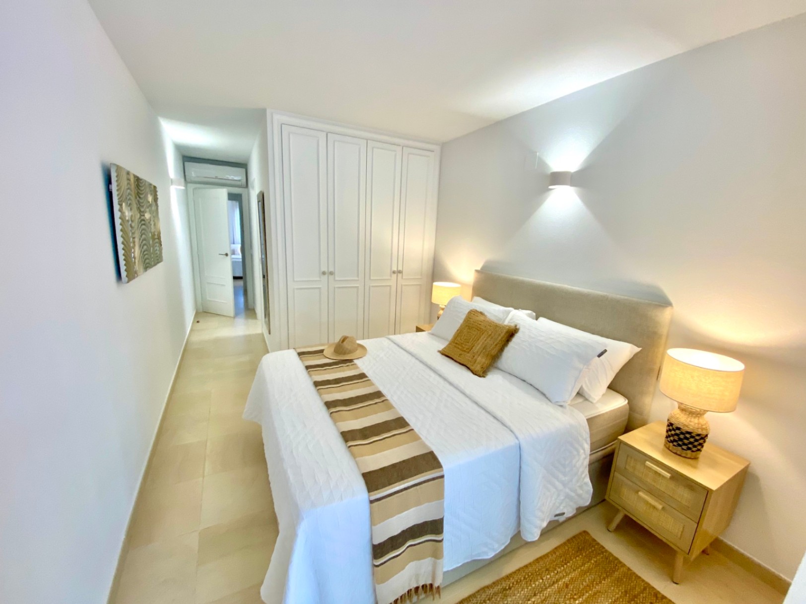 Dormitorio moderno con armarios empotrados blancos, cabecero tapizado en beige y mesitas de noche de madera con iluminación cálida sobre suelo cerámico.