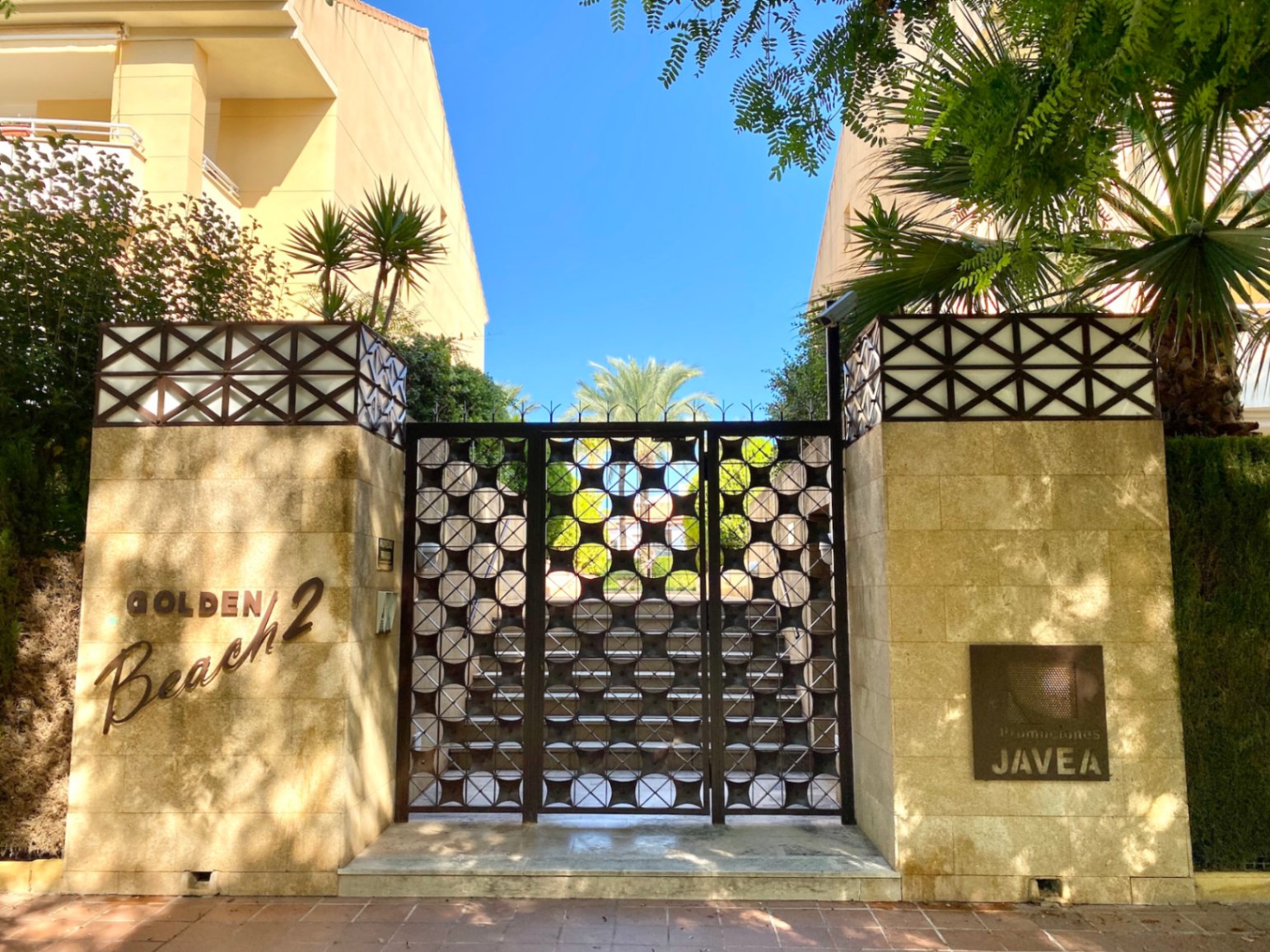 Entrada a Golden Beach 2 en Jávea, con puerta metálica de diseño circular entre pilares de piedra y vegetación mediterránea.