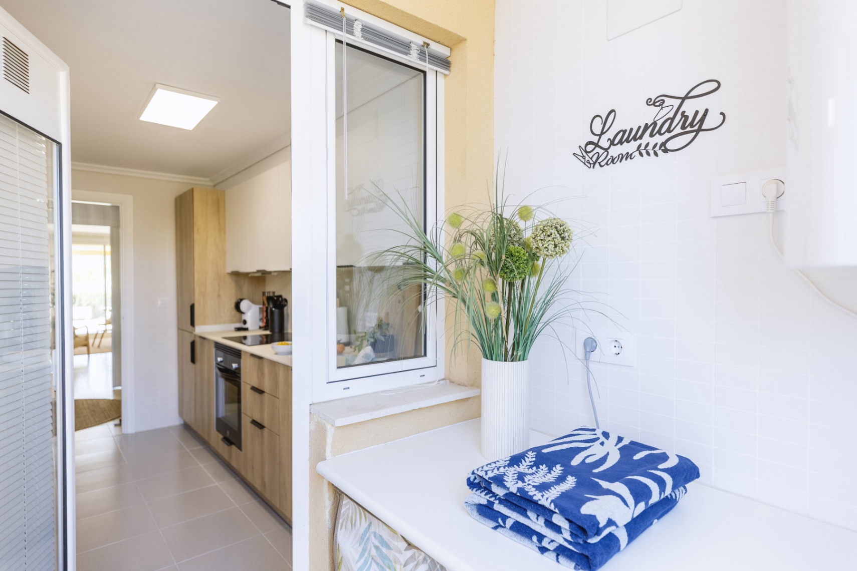 Lavadero funcional con encimera blanca y azulejos, situado junto a una cocina moderna con armarios de madera y electrodomésticos integrados.