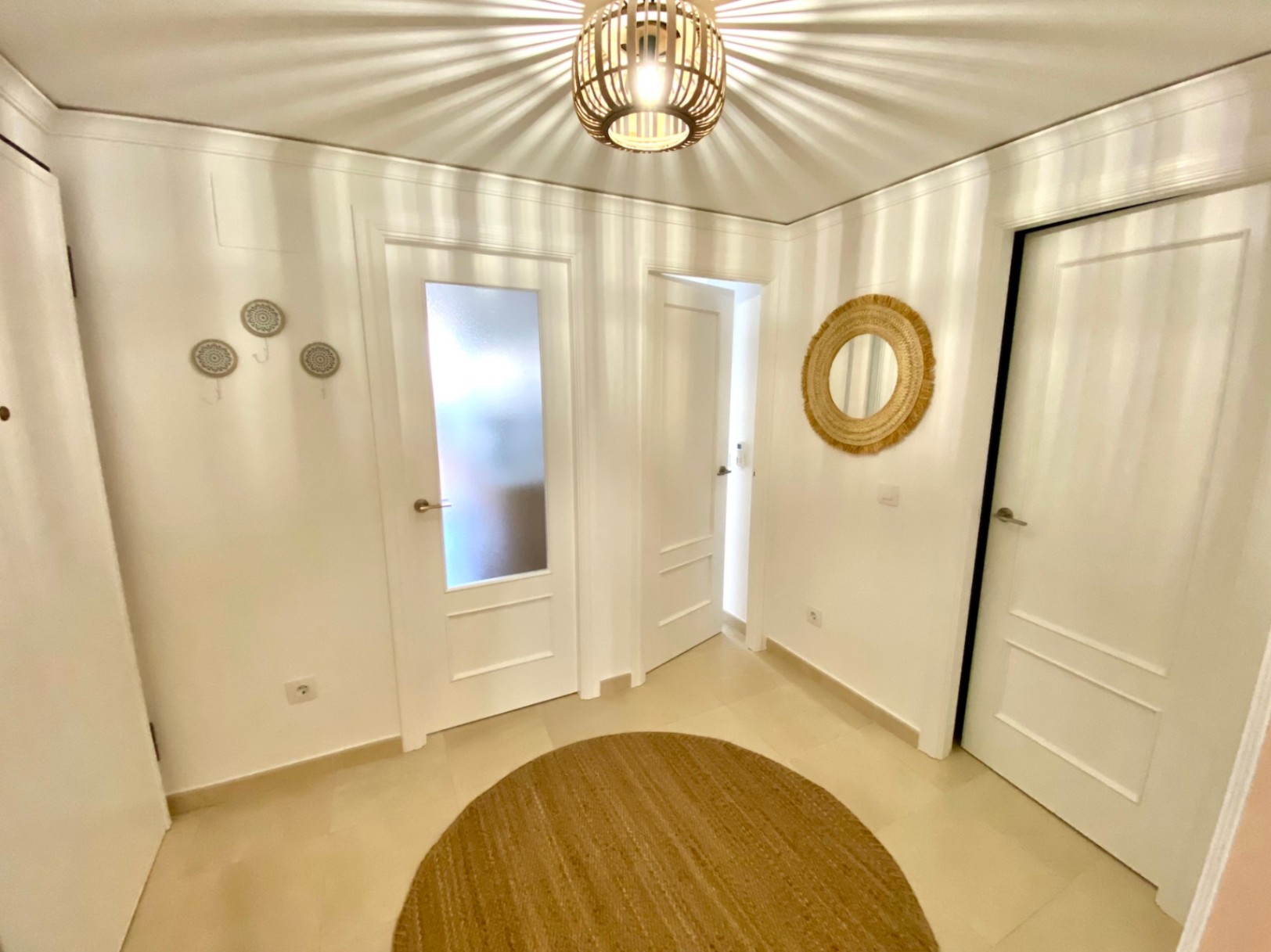 Pasillo con puertas blancas, alfombra redonda de yute y lámpara de techo de madera que proyecta sombras radiales en las paredes y el techo.