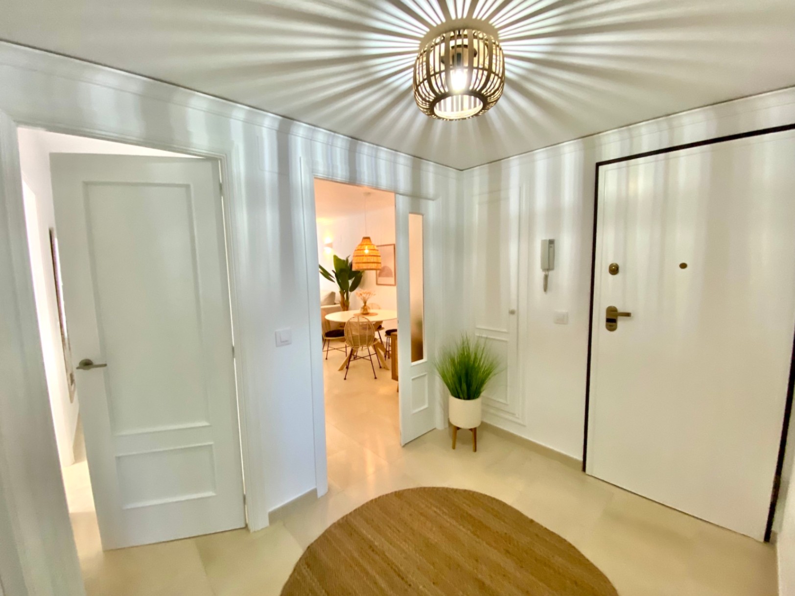 Recibidor luminoso con lámpara de techo de mimbre que proyecta sombras radiales, puertas blancas y vistas al comedor adyacente.