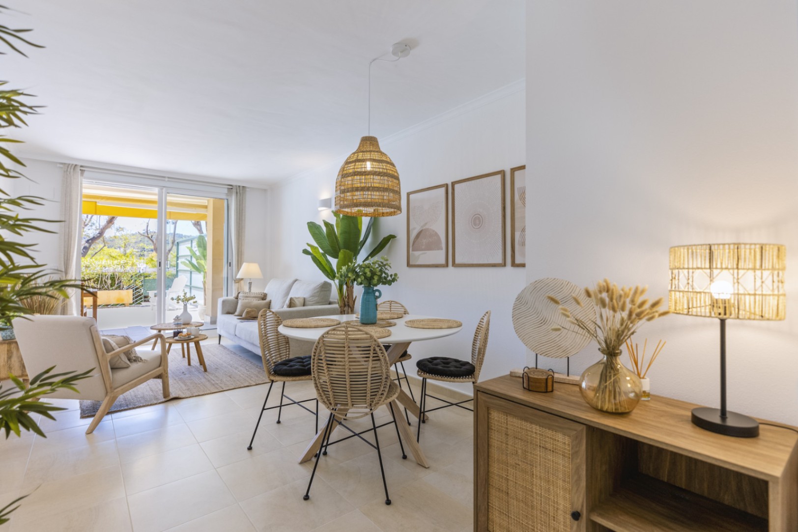 Salón-comedor luminoso con mesa redonda, sillas de mimbre y lámpara colgante de fibras naturales, conectado a una terraza exterior mediante puertas correderas.