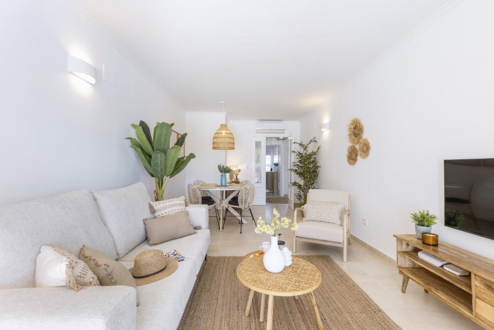 Salón-comedor luminoso con tonos neutros, sofá de tela, mesa de centro de mimbre y alfombra de fibras naturales bajo una iluminación de apliques cálidos.