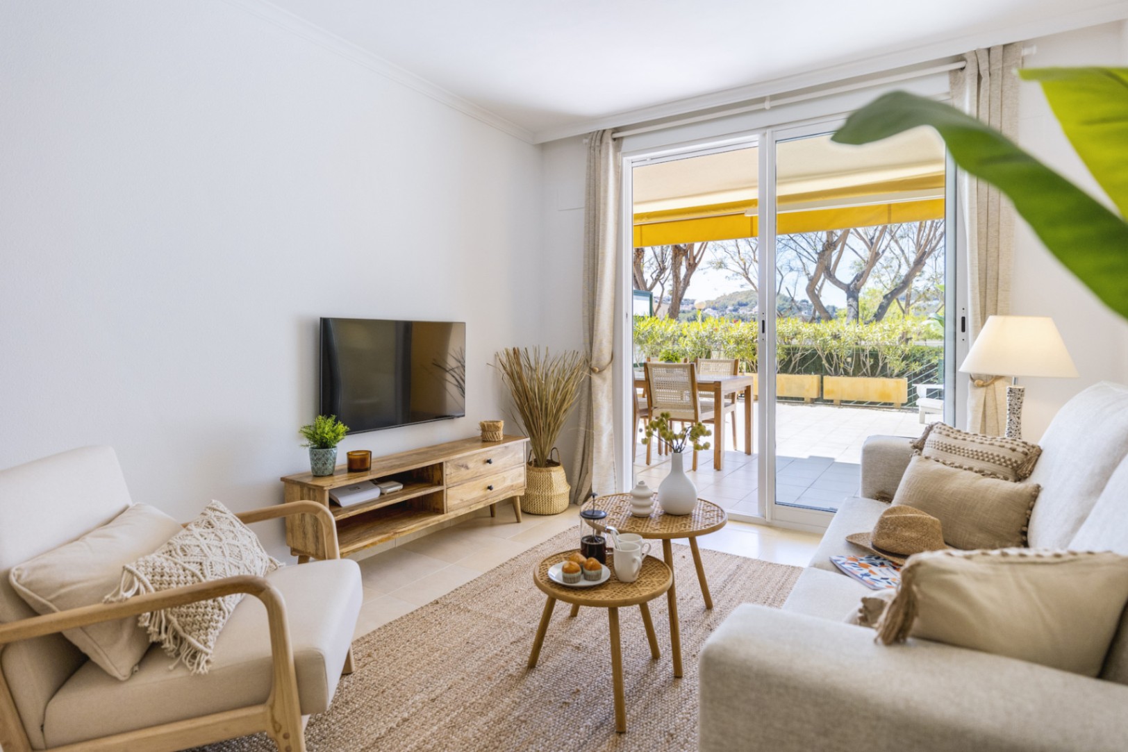 Salón luminoso con muebles de madera, alfombra de yute y mesas auxiliares de mimbre, conectado a una terraza exterior mediante grandes ventanales.