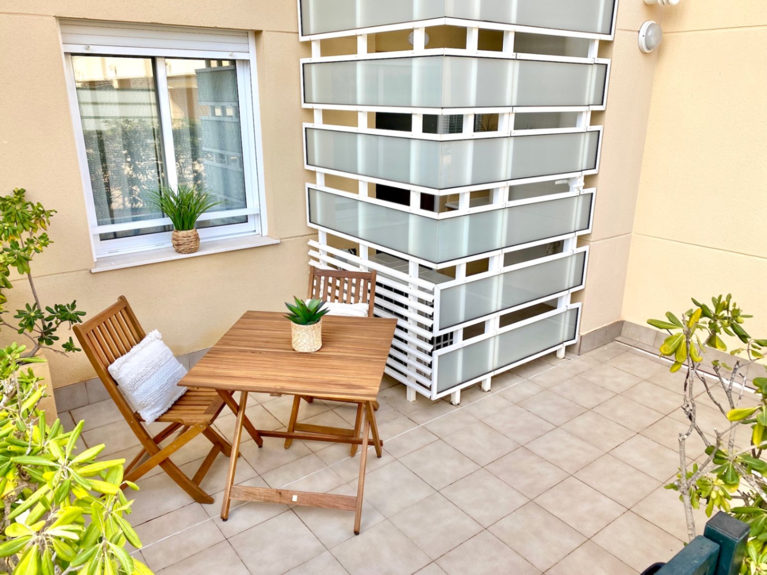 Terraza privada con suelo cerámico, conjunto de mesa y sillas plegables de madera y un panel divisorio de cristal traslúcido sobre paredes de tono arena.