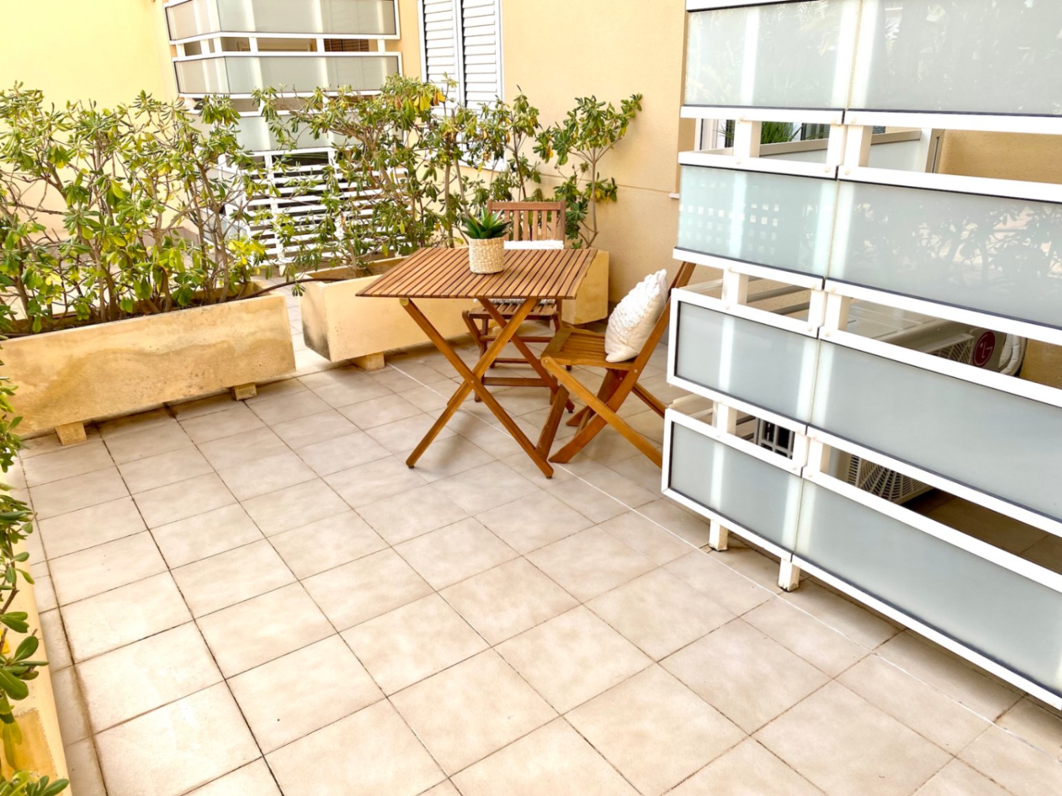 Terraza privada con suelo de baldosas beige, conjunto de mesa y sillas plegables de madera y mamparas de privacidad de vidrio esmerilado.
