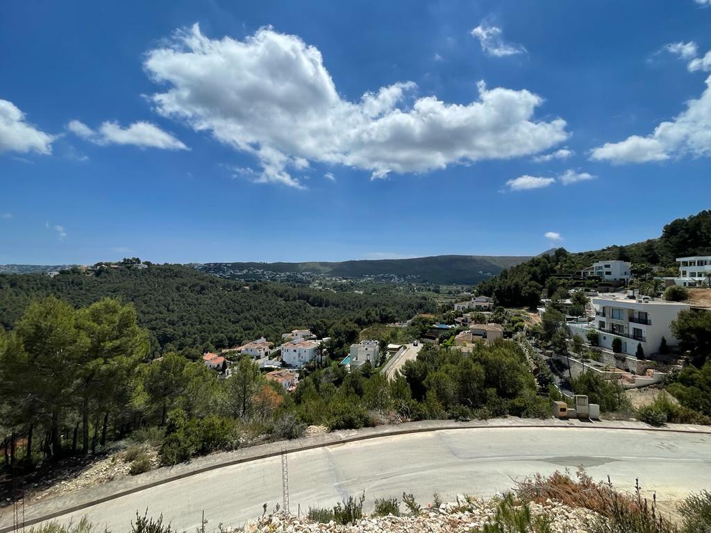 Vista panorámica elevada de un valle mediterráneo con villas blancas integradas en colinas de pinos, bajo un cielo azul brillante con nubes dispersas.
