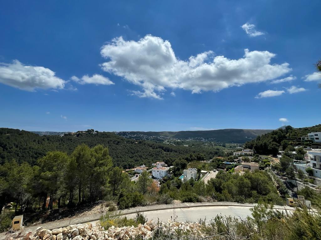 Vista panorámica elevada hacia un valle de pinos con villas blancas, colinas onduladas y un cielo despejado con nubes ligeras.