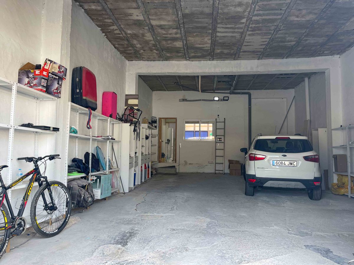 Amplio garaje con techos altos de hormigón visto y estanterías metálicas. Espacio funcional para vehículos y almacenamiento con entrada de luz natural al fondo.