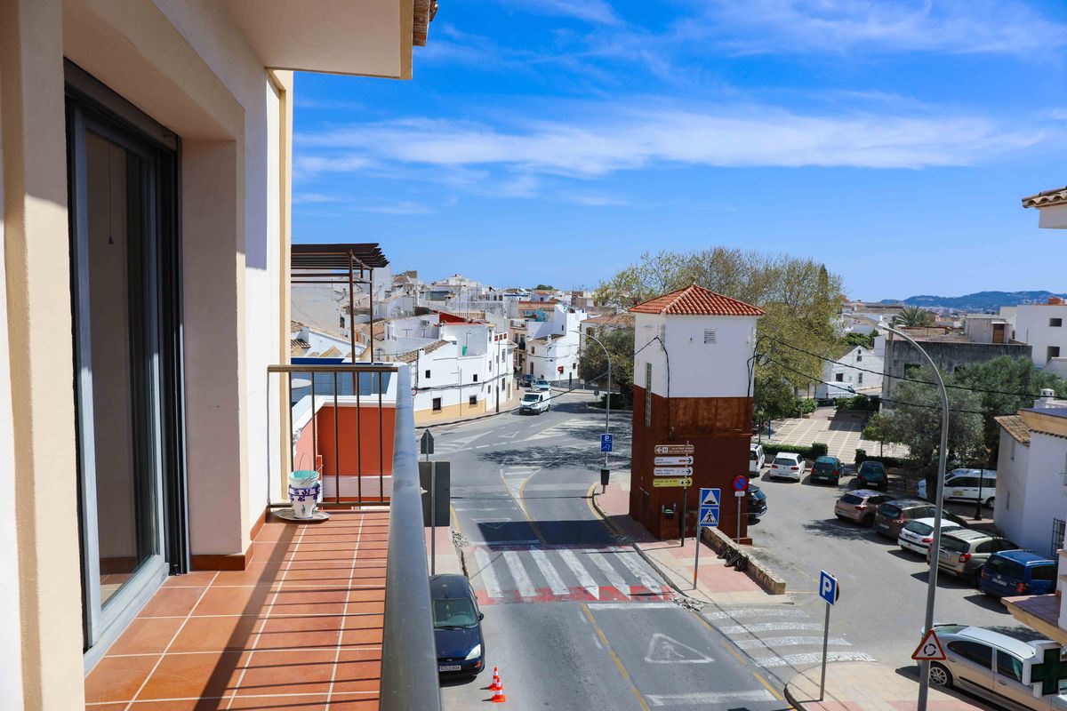 Balcón con suelo de baldosas de terracota y barandilla metálica con vistas a una intersección urbana y casas blancas de estilo mediterráneo.