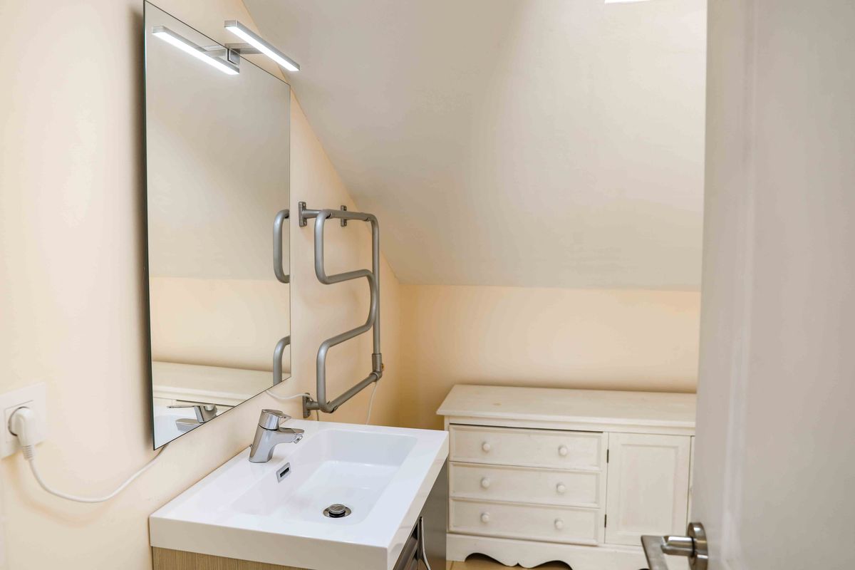 Baño abuhardillado con lavabo de cerámica blanca, espejo con iluminación LED integrada y toallero calefactable cromado bajo el techo inclinado.