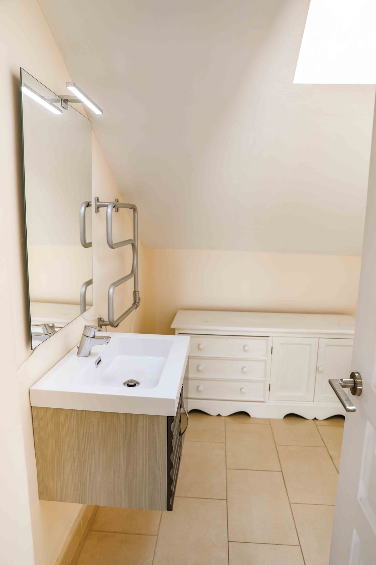 Baño abuhardillado con lavabo de cerámica blanca, mueble de madera, toallero térmico cromado y una cómoda blanca clásica sobre suelo cerámico beige.