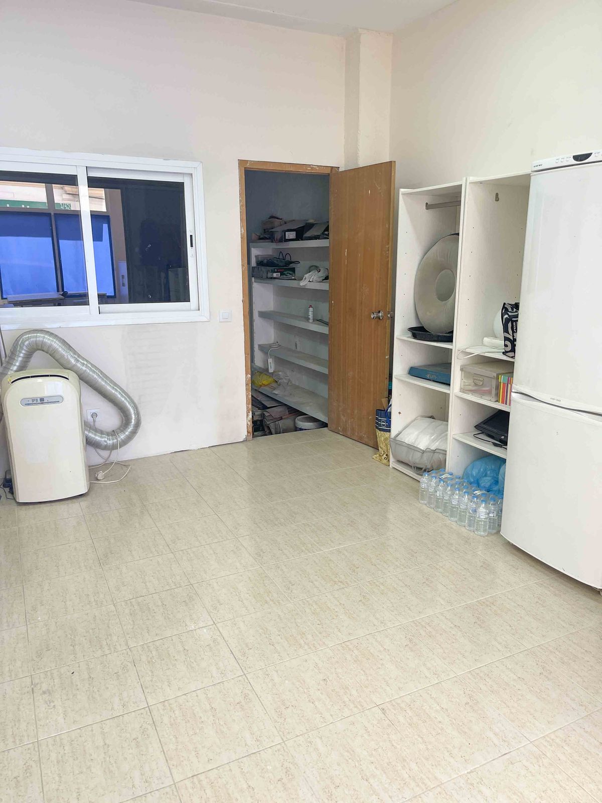 Cuarto de servicio con suelo cerámico claro, aire acondicionado portátil, estanterías blancas y despensa contigua con baldas integradas.