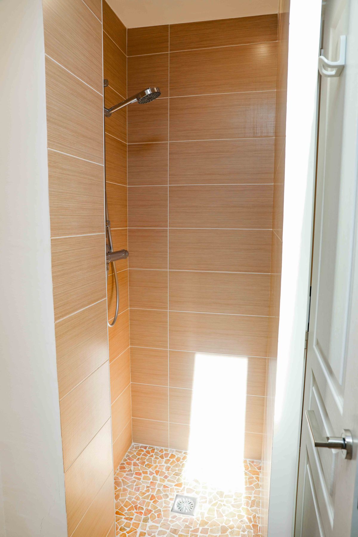 Ducha con azulejos cerámicos de efecto madera dispuestos horizontalmente y suelo de mosaico de cantos rodados. Incluye grifería cromada y entrada de luz natural.