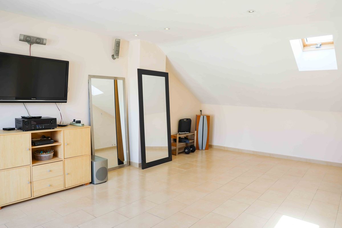 Habitación abuhardillada con techos inclinados, claraboya, suelo de baldosas claras y zona multimedia con televisor de pared y espejos de cuerpo entero.