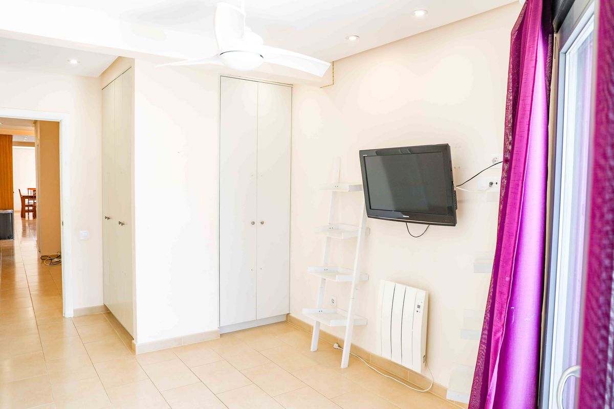 Habitación luminosa con armarios empotrados blancos, televisor de pared y estantería de escalera. Incluye ventilador de techo, focos empotrados y suelo cerámico.