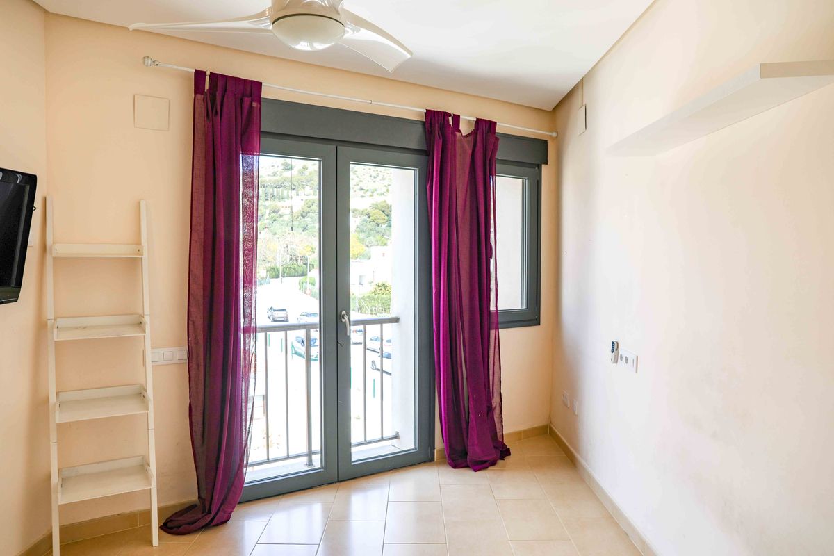 Habitación luminosa con puertas acristaladas de marco oscuro, cortinas púrpuras y ventilador de techo. Incluye una estantería blanca tipo escalera y suelos cerámicos claros.