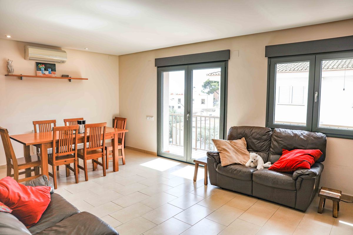 Salón-comedor luminoso con suelos cerámicos, conjunto de comedor de madera para seis comensales y sofá de cuero. Incluye acceso a balcón mediante puertas acristaladas.