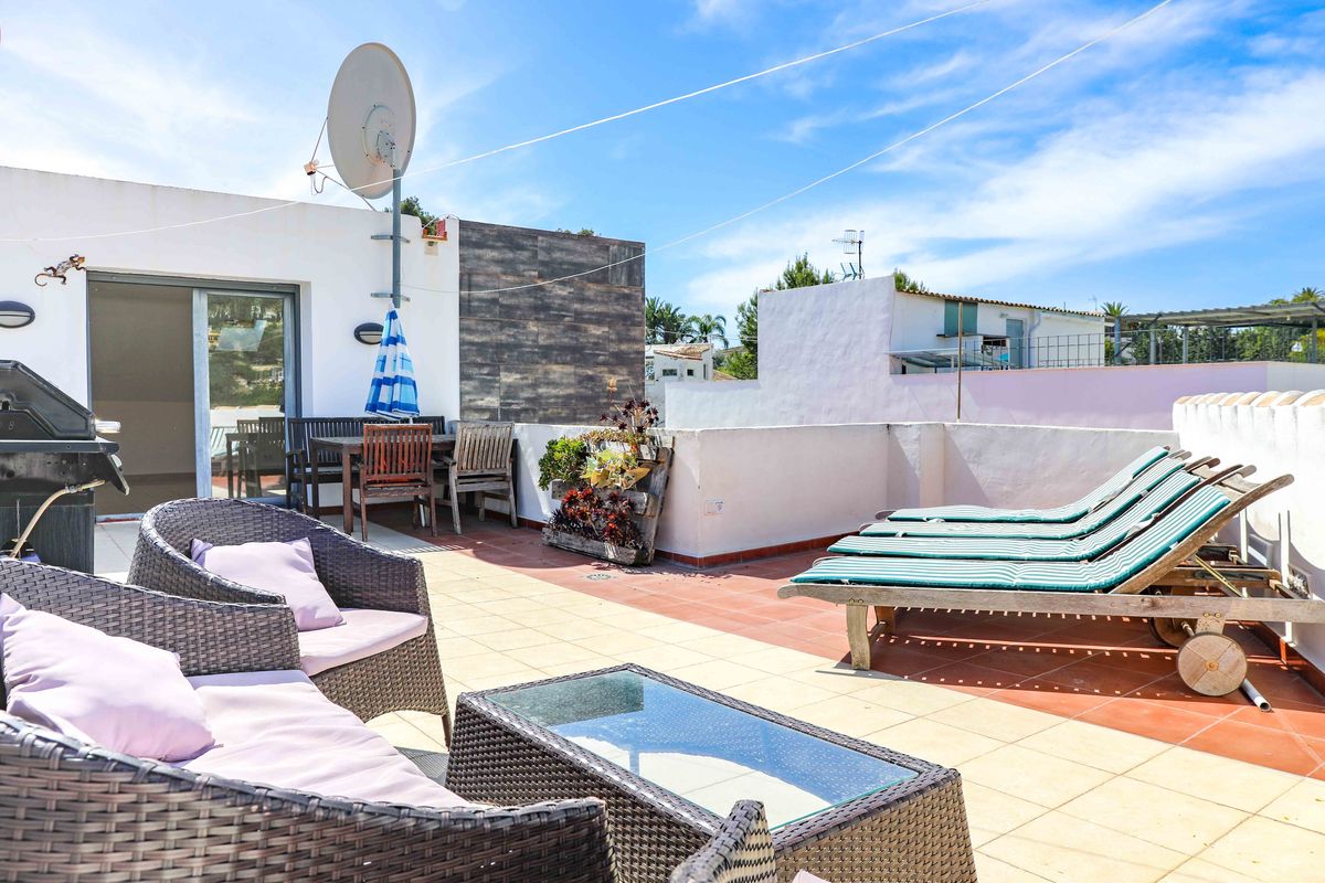 Terraza en la azotea con muebles de mimbre, tumbonas de madera y zona de comedor. Cuenta con suelos de baldosa, barbacoa y vistas despejadas bajo un cielo azul.