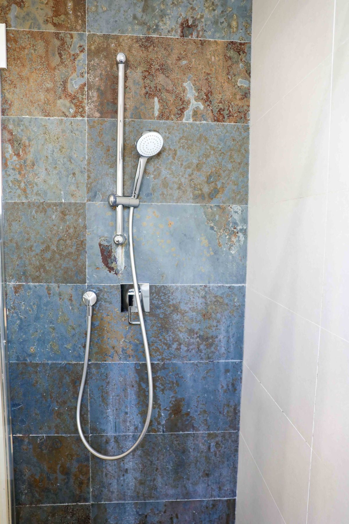 Zona de ducha con grifería cromada sobre riel deslizante y revestimiento de azulejos tipo pizarra en tonos óxido y azul, combinados con paredes claras.
