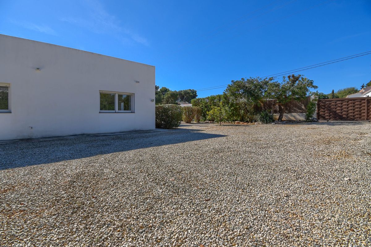 Amplia zona de aparcamiento de grava junto a una fachada blanca minimalista, con puerta de madera y vegetación perimetral bajo un cielo despejado.