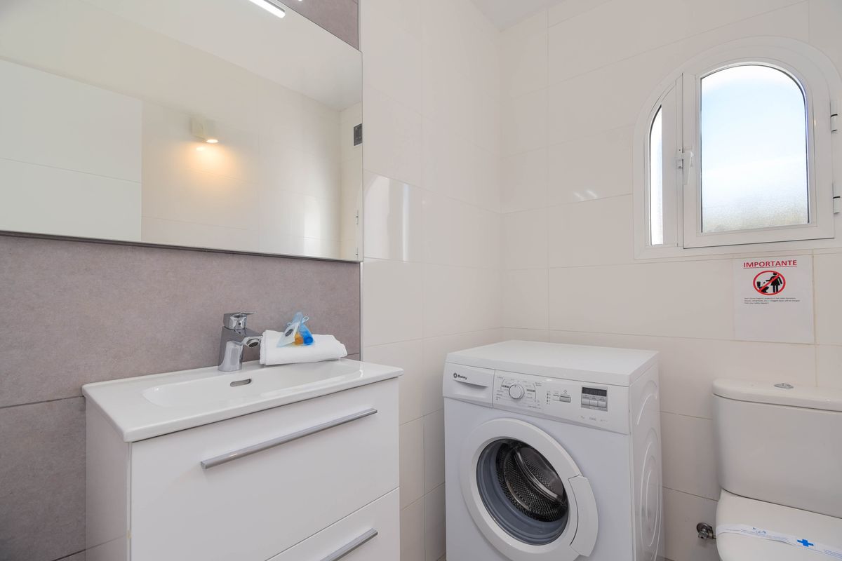 Baño funcional con mueble de lavabo blanco, espejo rectangular y lavadora integrada. La estancia cuenta con paredes alicatadas y una ventana de arco con cristal mate.