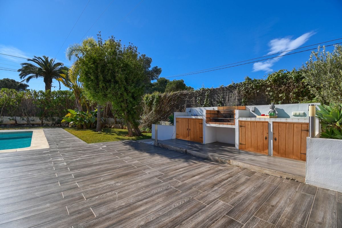 Cocina exterior de obra con barbacoa de ladrillo, armarios de madera y fregadero integrado sobre una terraza de gres porcelánico junto a la piscina.