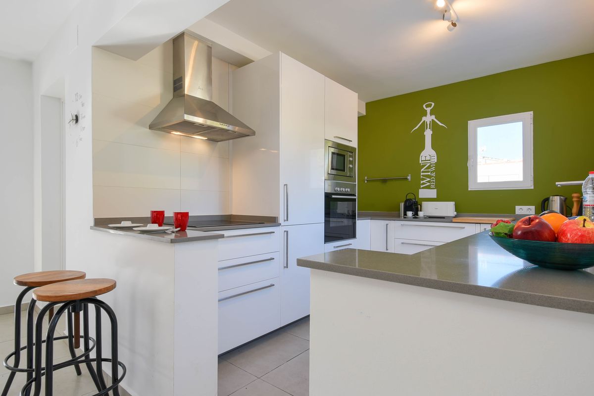 Cocina moderna con mobiliario blanco, encimeras grises y pared de acento verde. Incluye electrodomésticos integrados, barra de desayuno con taburetes y campana de acero.
