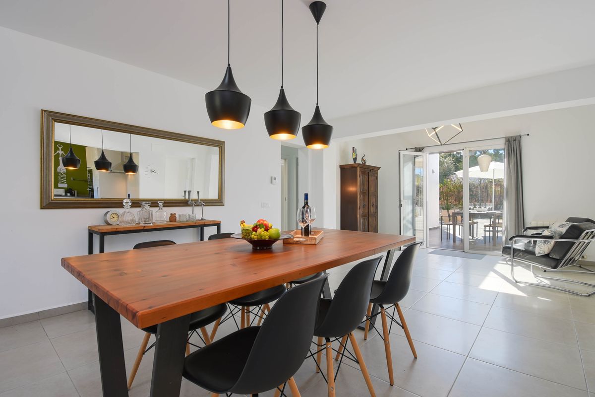 Comedor moderno con mesa de madera maciza, sillas de diseño negras y lámparas colgantes, conectado directamente a una terraza exterior soleada.