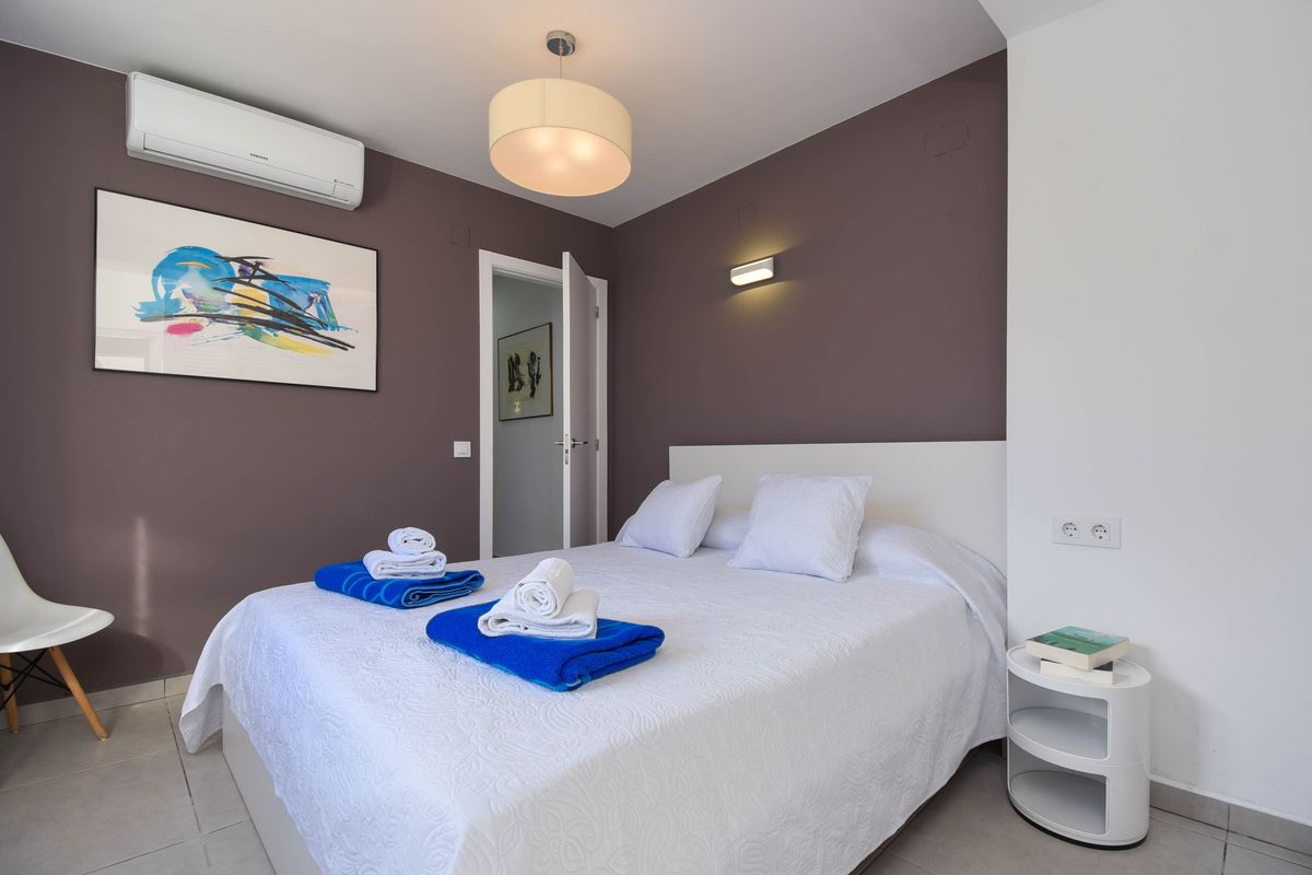 Dormitorio moderno con pared de acento malva, arte abstracto y cama doble con textiles blancos. Incluye aire acondicionado y una mesita de noche circular de diseño.