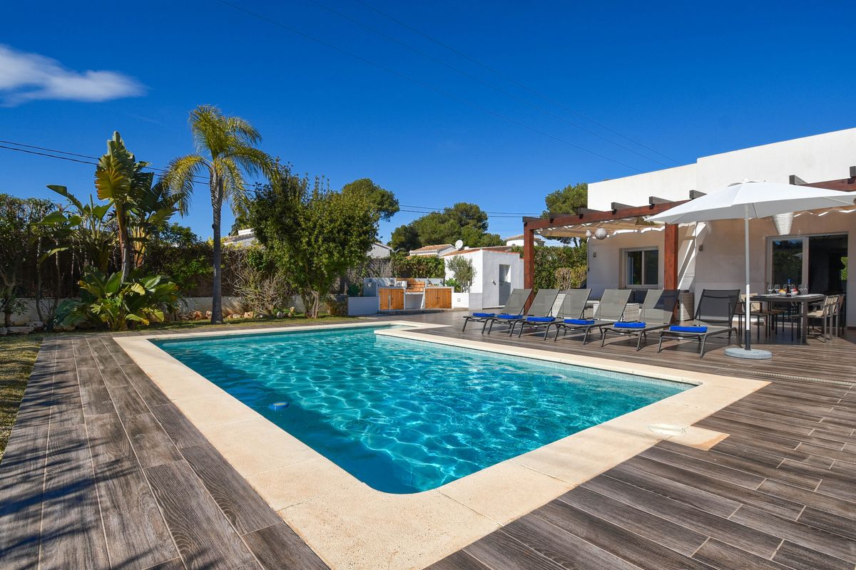 Piscina exterior con borde de piedra, terraza de gres efecto madera, tumbonas y zona de comedor bajo una pérgola de madera.