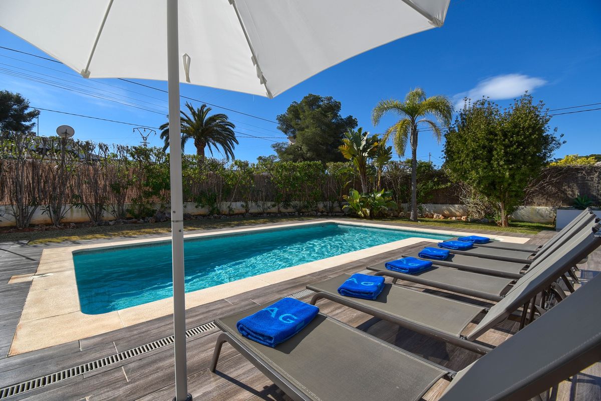 Piscina rectangular exterior con borde de piedra, terraza de gres porcelánico efecto madera, tumbonas grises con toallas azules y vegetación mediterránea.