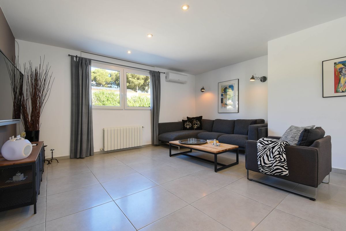 Salón moderno con sofá rinconero gris, mesa de centro de madera y suelos cerámicos. Dispone de aire acondicionado, calefacción y vistas al jardín exterior.