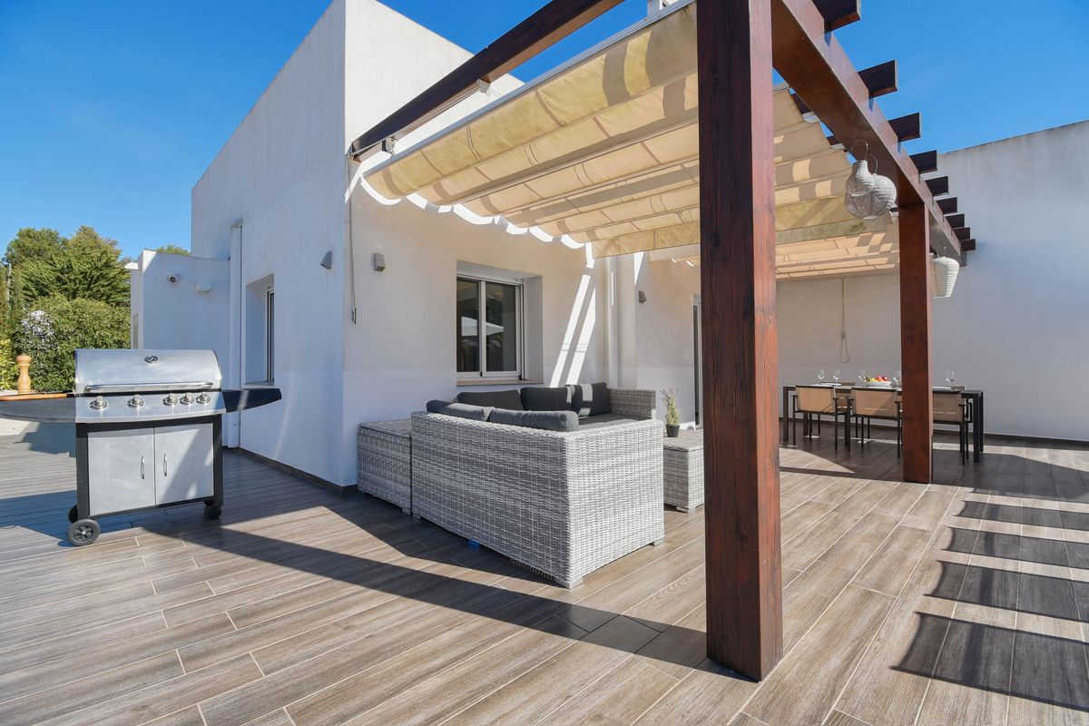 Terraza exterior con suelo cerámico imitación madera, pérgola de vigas oscuras con toldo retráctil, zona de estar de mimbre gris y barbacoa de acero inoxidable.
