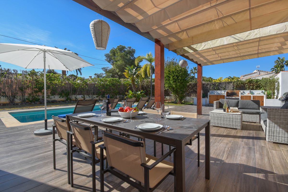 Terraza exterior con zona de comedor bajo toldo retráctil, suelo cerámico imitación madera, piscina y área de descanso con cocina de verano al fondo.
